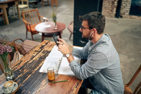 Mladý Muž Sedí Sám Kavárně Používá Chytrý Telefon Poslouchá Sluchátka — Stock fotografie