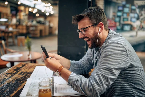 快乐的年轻商人 带着眼镜和耳机 在咖啡店里用智能手机 一个人坐在桌旁 现代内部背景 — 图库照片