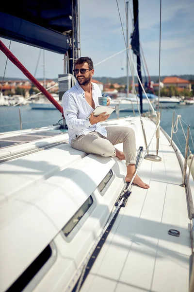 タブレットを持っている若い男がヨットに座っていて 海辺の晴れた日にドックのコーヒーと美しい景色を楽しんでいます — ストック写真