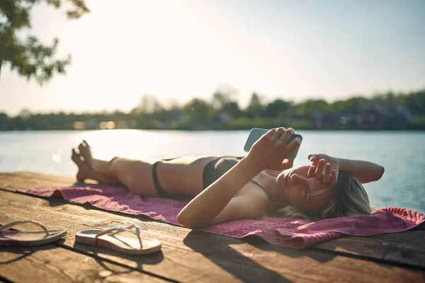 夏天阳光明媚的日子里 穿着比基尼 拿着手机站在河边码头上的年轻美女 — 图库照片