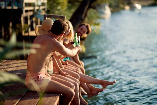 四个年轻朋友一起度假 坐在水边的木制防波堤上 喝着饮料欢呼 生活方式的概念 — 图库照片