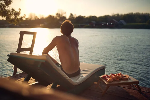 英俊的年轻人坐在日光浴床上看着水 暑假时人们的后视镜 生活方式的概念 — 图库照片