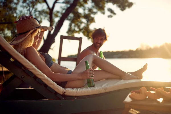 水着を着た若いカップルが日向ぼっこをして 美しい夏の日に川の岸で太陽と飲み物を楽しんでいます — ストック写真