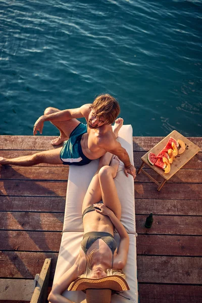 青い水でカップル日光浴の高角度ショット 木製の桟橋に フルーツプレートと彼らの側で飲み物と ライフスタイルの概念 — ストック写真