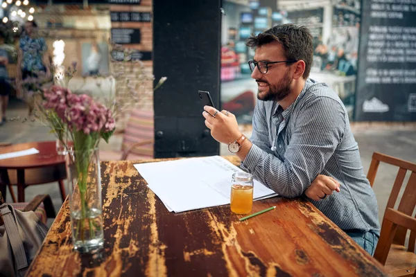 一个戴眼镜的英俊年轻人用智能手机 在现代咖啡馆里喝冷饮的人 — 图库照片