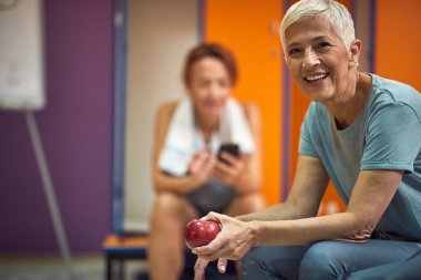 Spor giyim bölümünde kıdemli bir kadın. Gülümsüyor ve soyunma odasında oturuyor. Elma tutuyor, formda ve sağlıklı.. 