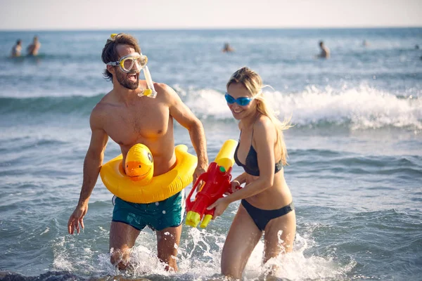 ビーチで水鉄砲で陽気な若いカップル ゴム製のアヒルのプールが浮かぶ男 夏休みのコンセプト — ストック写真