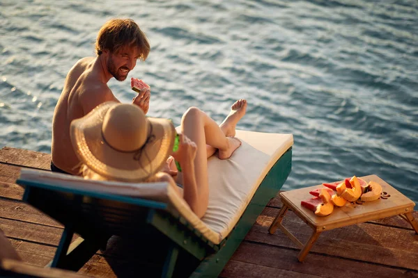 夫妇日光浴在木制防波堤和吃西瓜 戴着草帽的女人躺在日光浴床上 生活方式概念 — 图库照片