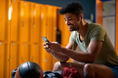 Kulaklıklı genç sporcu, soyunma odasında akıllı telefon kullanarak antrenmana hazırlanıyor. Spor, yaşam tarzı, sağlık konsepti.