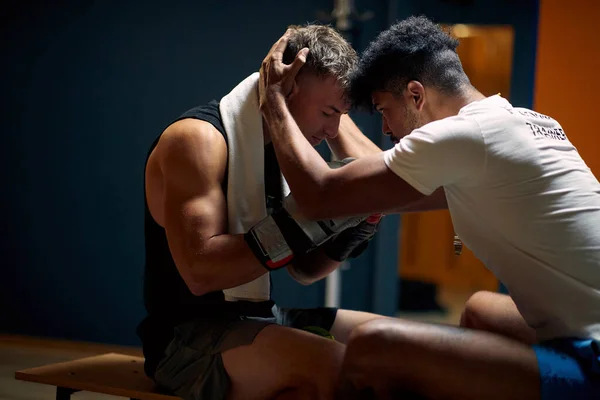 Coach helping young boxing player mentally prepare, holding his head and talking to him, in dressing room sitting on bench. Sports, martial arts, lifestyle concept.