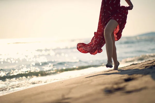 若い女性はビーチの海岸を歩いている 赤いドレスの女性は 足に焦点を当てます ファッション ライフスタイルのコンセプト — ストック写真