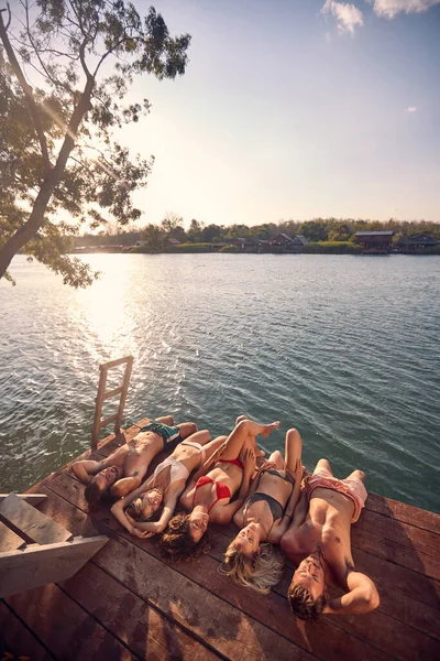 水着で4人の友人は 水の日光浴で木製の桟橋に横たわっていた 休暇で若い男と女 ライフスタイルの概念 — ストック写真