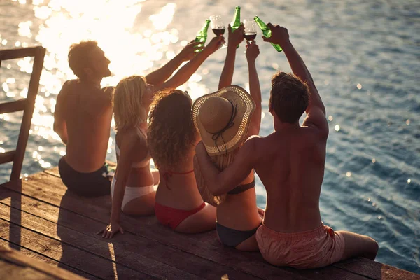 水で木製の桟橋に座っている若者のグループの背面ビューショットでは 飲み物で応援 休暇中のカップル ライフスタイルの概念 — ストック写真