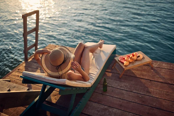 若い女性は水で木製の桟橋で日光浴を楽しんでいます 彼女の大きなファッショナブルなわらの帽子をポーズと保持 ライフスタイルのコンセプト — ストック写真