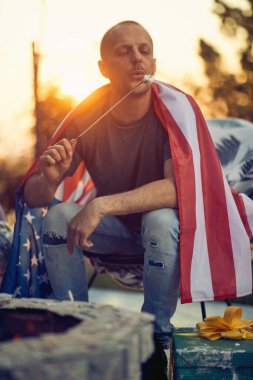 Orta yaşlı bir adam, omuzlarında ABD bayrağı ile bir sandalyede oturuyor, açık havada, günbatımı arka planında, sıcak bir kulübede ızgara marşmelovların tadını çıkarıyor. 4 Temmuz Bağımsızlık Günü