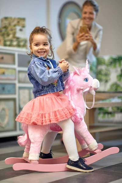可爱的小女孩骑着粉红独角兽在商店里摇着马 高兴极了 妈妈在后面拍照 生活方式概念 — 图库照片