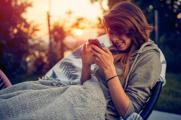 絵のように美しい屋外の環境に座っている間 女の子は夕日の魅惑的な魅力に魅了されています 携帯電話を手に 彼女は純粋な喜びの瞬間に没頭している — ストック写真