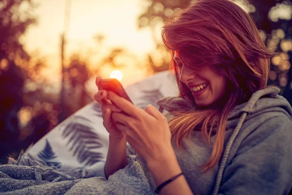 女の子は屋外に座って 彼女の顔は輝く笑顔で照らされながら 彼女の携帯電話に包まれています 美しい夕日の暖かい色合いに包まれた穏やかな裏庭です — ストック写真