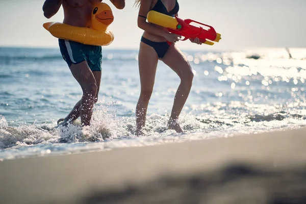 幸せな若い男性と女性とのクレイジーな休暇やビーチでの水銃で楽しい 若いカップルは夏休みにビーチで楽しみを持っています — ストック写真
