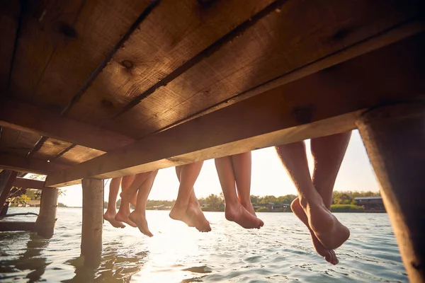 若者の足に焦点を当てます 4人の若い友人が水のそばに座っている 足の後ろのビュー 楽しさ ライフスタイルの概念 — ストック写真