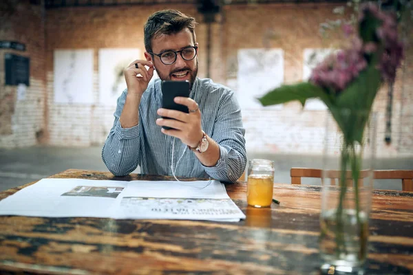 年轻迷人的男人坐在咖啡馆里 带着耳机用的是智能手机 现代咖啡馆背景 — 图库照片