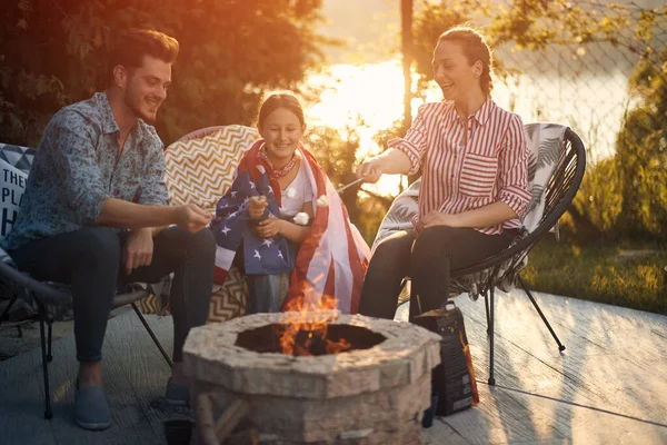 stock image A family gathers around a crackling fire, joyfully roasting marshmallows as they celebrate Independence Day. Little girl wrapped in the embrace of an American flag, her eyes sparkling with excitement.