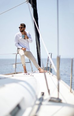 Genç ve yakışıklı bir erkek model, deniz kenarındaki bir yatta fotoğraf çekiminde güneşli bir günde kahve ve güneşin tadını çıkarıyor. Yaz, deniz, tatil