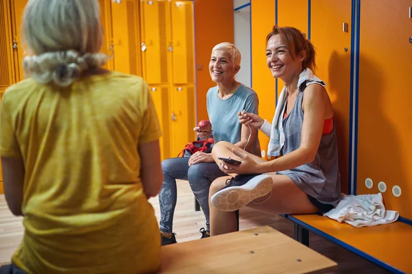 与年轻女教练一起坐在体育馆更衣室里的老年妇女在锻炼后喋喋不休 生活方式概念 — 图库照片