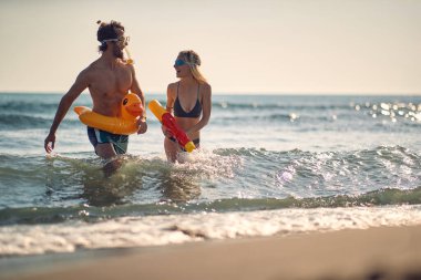 Kumsalda genç ve çekici bir çift. Su tabancalı bir kadın, plastik ördek havuzlu bir adam. Tatil, yaz tatili, yaşam tarzı konsepti.