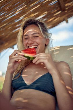 Kumsalda karpuz meyvesi yiyen, güneşlenen genç sarışın kadın. Yaz tatili. Tatil, cüzzam, yaşam tarzı konsepti.