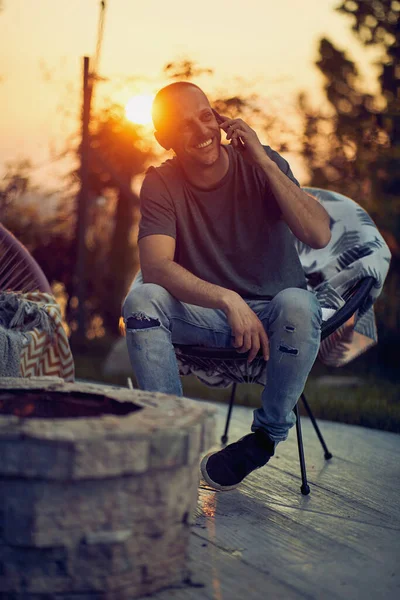一个快乐的中年男人坐在茅屋里舒适的椅子上 在电话里聊天 身后是日落 生活方式概念 — 图库照片