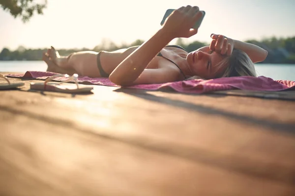 迷人的健康的成年女子躺在木制防水堤上晒太阳 看她的手机 生活方式概念 — 图库照片