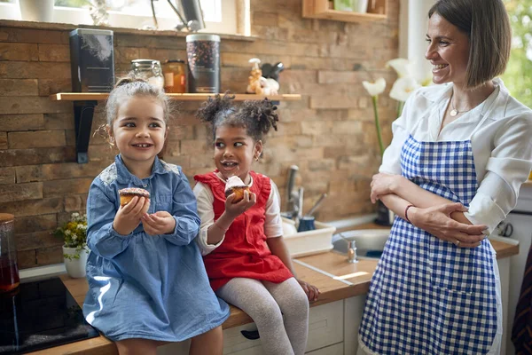 两个姑娘 一个皮肤白皙 另一个肤色发亮 肩并肩坐在厨房里 品尝着美味的松饼 站在她们旁边 她们的母亲骄傲地 爱心地笑着 — 图库照片