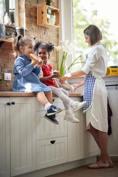 两个可爱的小女孩坐在柜台顶上吃着小吃 西瓜和松饼 旁边的妈妈在洗碗 生活方式概念 — 图库照片