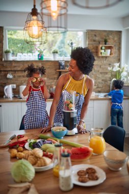 Anne ve kızı birlikte mutfağa gelip sağlıklı bir yemek hazırlıyorlar. Sevimli kız, şakacı bir şekilde gözlerini salatalık dilimleriyle kapatıyor. Arka planda, kızın kardeşinin bulaşıkları yıkadığını görüyoruz..