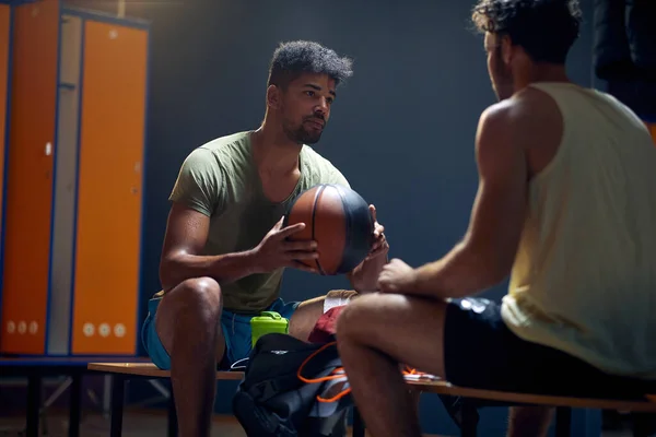 两个年轻英俊的篮球运动员坐在长椅上的更衣室里 彼此交谈着 锻炼之后 积极生活方式概念 — 图库照片