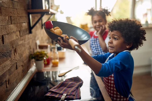 非洲裔美国人的兄弟姐妹在厨房 那个年轻的女孩 站在后面 在前景展望中 她哥哥举起一个盛满蔬菜的热腾腾的煎锅 — 图库照片