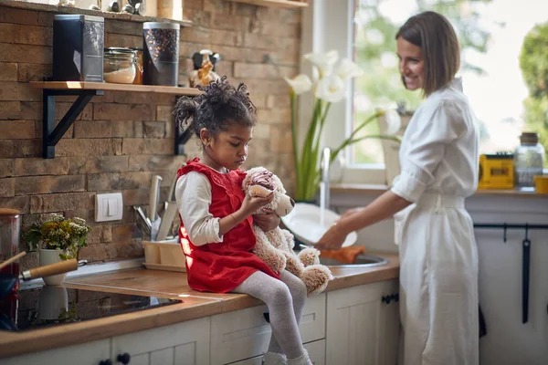 小女孩一只手拿着心爱的泰迪熊玩具 一边享用着美味的松饼 在她身边站着她慈爱的母亲 勤勤恳恳地洗碗 带着呵护的微笑 — 图库照片