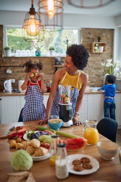 妈妈和女儿一起在厨房里准备健康的饭菜 可爱的女孩开玩笑地把黄瓜片放在她的眼睛上 在背景中 我们看到女孩的哥哥在洗盘子 — 图库照片