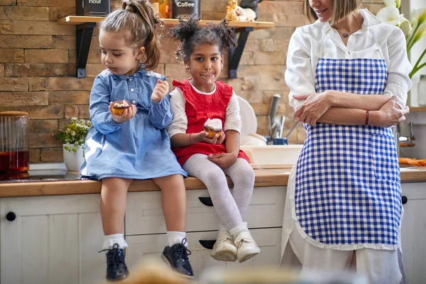 二人の若い女の子は 彼らがおいしいカップケーキマフィンを保存し キッチンカウンターに座って楽しい瞬間にキャプチャされます 純粋な喜びと期待の笑顔で 彼らはカップケーキを保持します — ストック写真