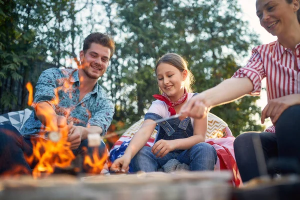 家族は裏庭に集まり 一緒に楽しむことができます 両親と彼らの娘は 激しい火災の上にマシュマロを焙煎するという単純な喜びにふけるのを見ています — ストック写真