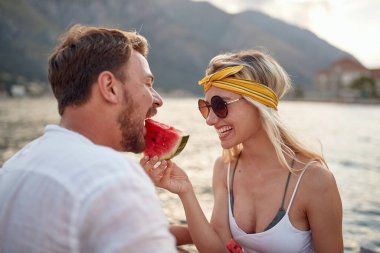 Genç bir çiftin güzel bir günde rıhtımda otururken birbirlerini sulu bir karpuzla beslemeleri. Aşk, ilişki, tatil, deniz