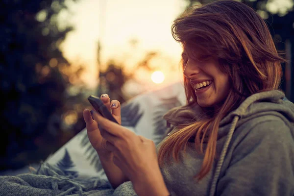 スマートフォンで美しい喜びの若い女性のポートショットを閉じます 屋外で彼女の時間を楽しんで 居心地の良い椅子に座って 日没の背景 — ストック写真