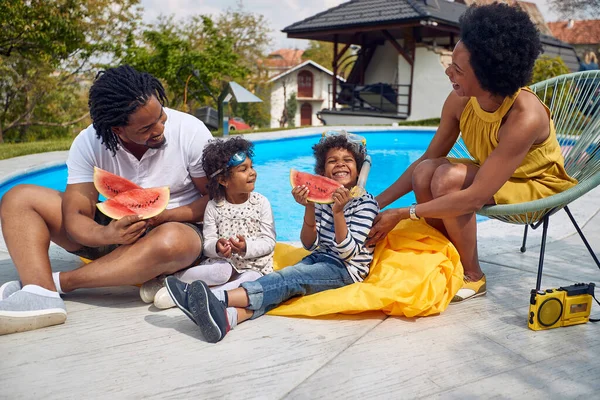 Summer moments of an Afro-American family by the poolside. The parents, along with their children, a boy and a girl, come together to savor the delights of a refreshing watermelon feast.