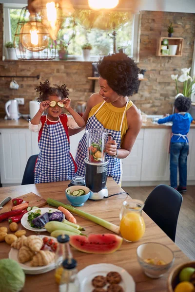 妈妈和女儿一起在厨房里准备健康的饭菜 可爱的女孩开玩笑地把黄瓜片放在她的眼睛上 在背景中 我们看到女孩的哥哥在洗盘子 — 图库照片