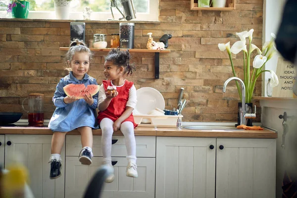 可爱的姑娘们坐在厨房的台子上 吃着下午的小吃 西瓜和松饼 在一个现代化的厨房里 她们在一起感到很快乐 生活方式概念 — 图库照片