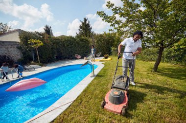 Aile havuz kenarında, Afro-Amerikan adam çim biçiyor, yaz aktivitelerine hazırlanıyor. Aile bir arada.