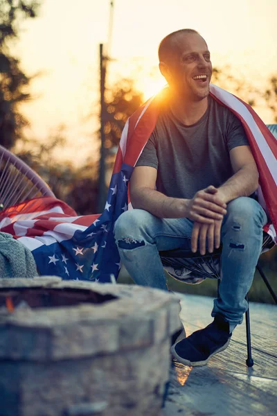 Full body vertical shot of cheerful man with the flag of USA on his back, relaxing in a cozy chair outdoors. 4 fourth July Independence Day
