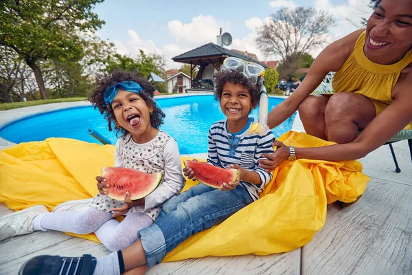 Afro-American family by the poolside. The mother and her children, a boy and a girl, sit together basking in the warm sunshine. The children delightfully indulge in juicy slices of watermelon.