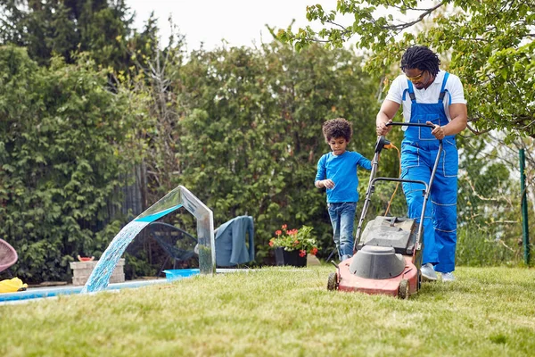 若い父親は屋外の電気芝刈り機で草を刈り 息子は彼を助けている チーム 息子として一緒に働く ホーム ライフスタイル ガーデニングコンセプト — ストック写真